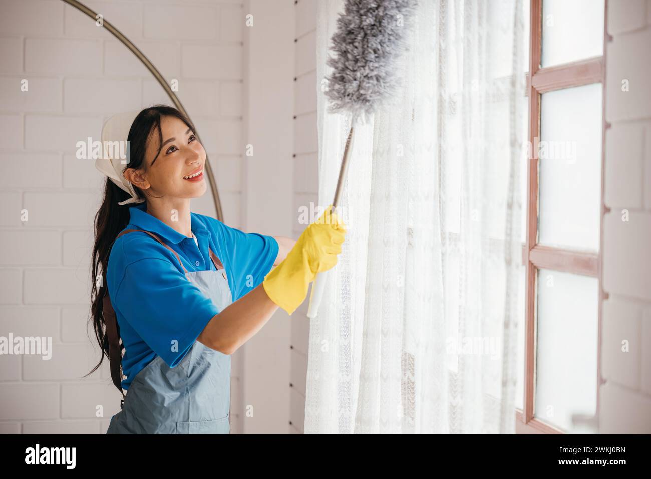 Occupation in action, Asian woman dusts window blinds holding duster. Her smile shows happiness in Stock Photo