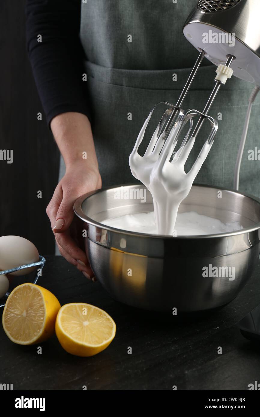 Woman making whipped cream with hand mixer at black table, closeup