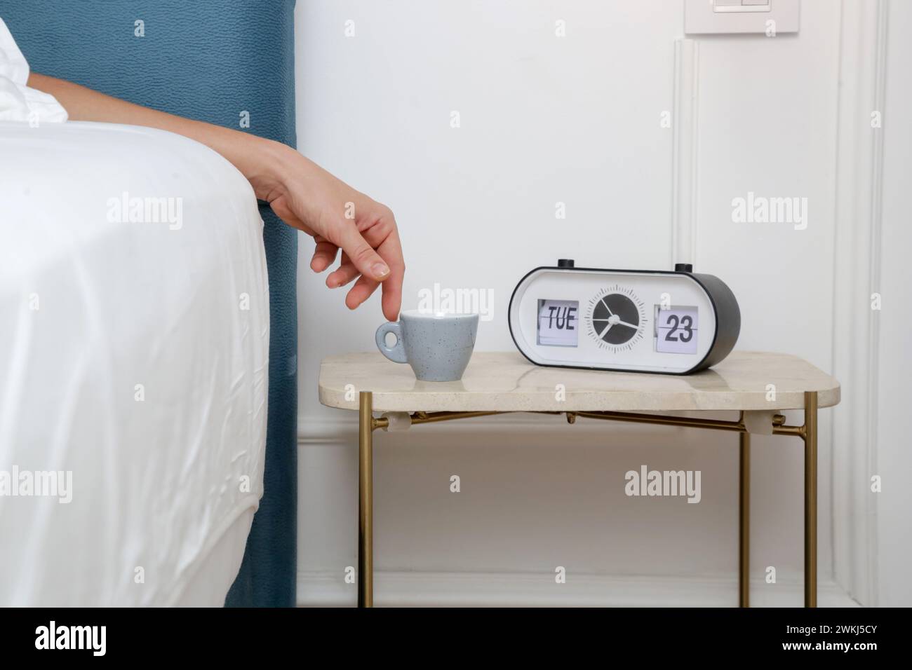 Woman in bed reaching for her first morning coffee from the nightstand. A light blue espresso cup on the bedside table next to the alarm clock Stock Photo