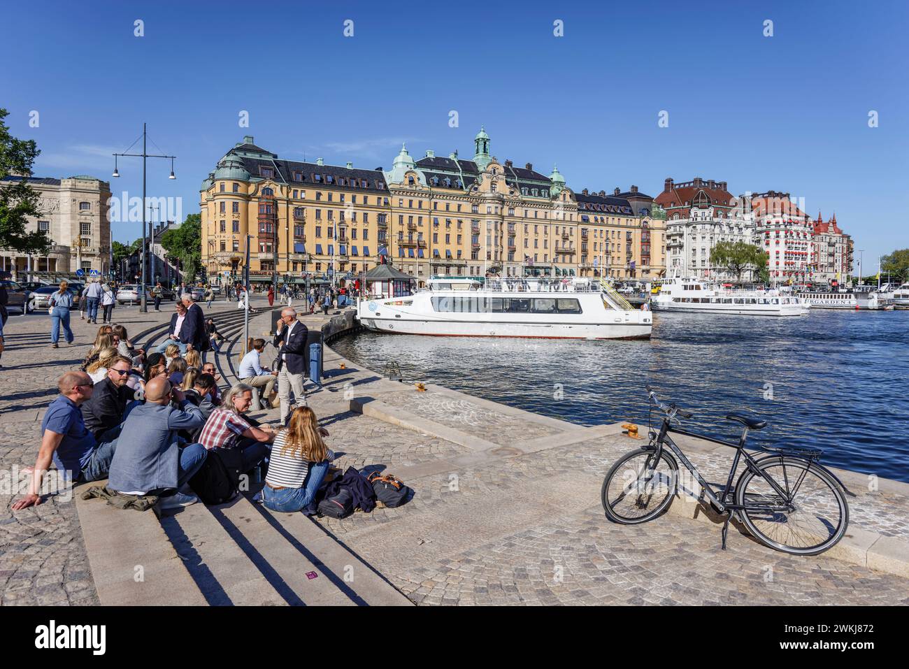 People relax on waterfront by Nybroviken at Strandvagen with Kvarteret Bodarne art nouveau building and Svenskt Tenn behind. Ostermalm Stockholm Stock Photo