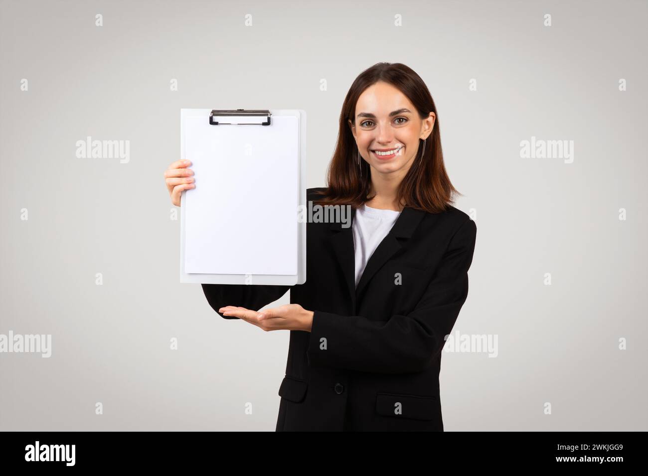 A professional young woman in a black suit confidently presents a blank white clipboard with a smile Stock Photo