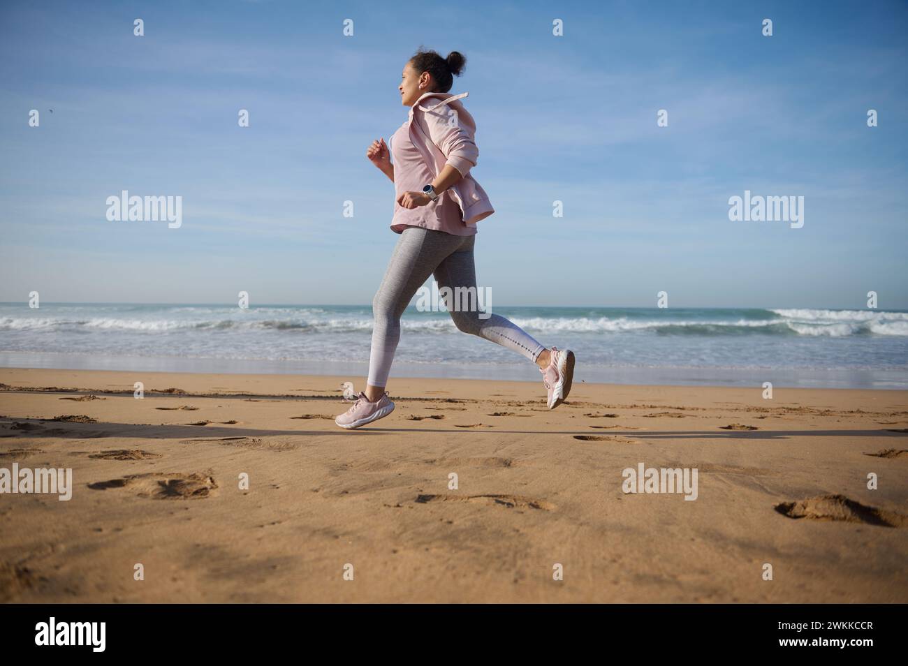 Full length side portrait of a sportive woman exercising outdoor, running, jogging on a sandy beach on warm sunny day. Athletic female runs along the Stock Photo