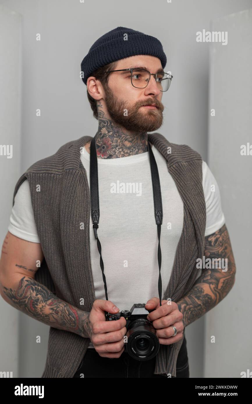 Brutal handsome professional photographer hipster man with glasses with tattoos with a knitted hat and sweater holds a photo camera and stands in the Stock Photo