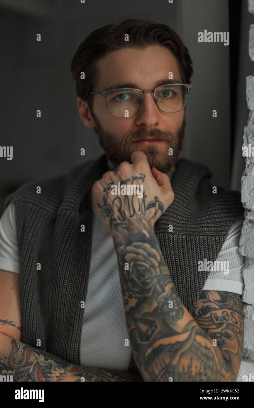Stylish handsome fashionable hipster man with a hairstyle and beard with a tattoo with fashion glasses looks at the camera in the room Stock Photo