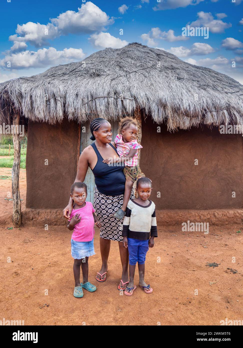 Happy village african family, mother with braided hair together with the three kids in front of the mud house with thatched roof Stock Photo