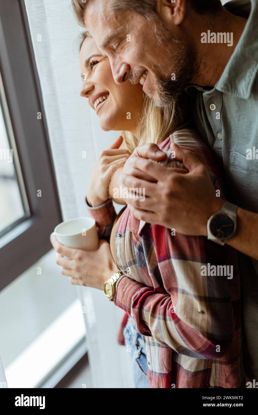 In a softly lit room, a man lovingly embraces a woman from behind as they both gaze out the window, sharing a quiet, intimate moment together Stock Photo