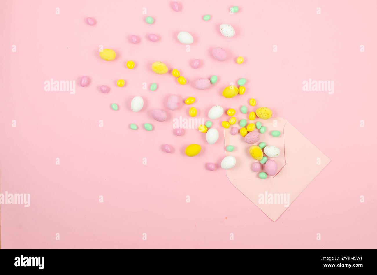 Empty white card in envelope and scattered chocolate Easter colored candy eggs on pink background. Easter concept. Stock Photo