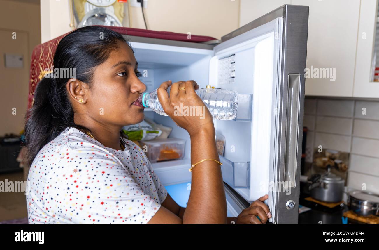 A South Asian woman takes a moment to refresh with a sip of water from a reusable bottle in her warm and inviting kitchen Stock Photo