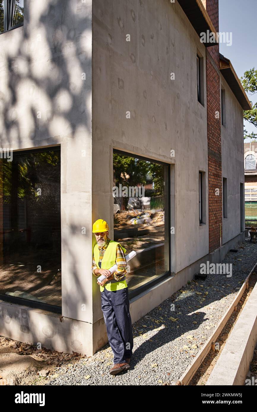 good looking dedicated cottage builder in safety gear with blueprint and posing on construction site Stock Photo