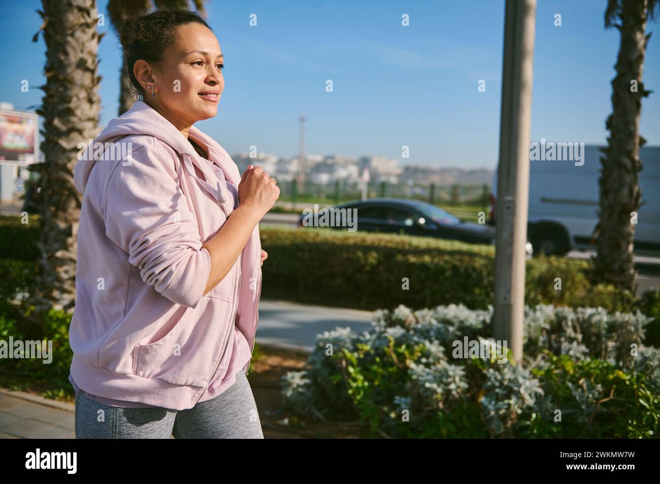 Beautiful young adult woman running on the city park in the morning. Fitness woman burning calories during morning jog, enjoying her workout outdoor. Stock Photo
