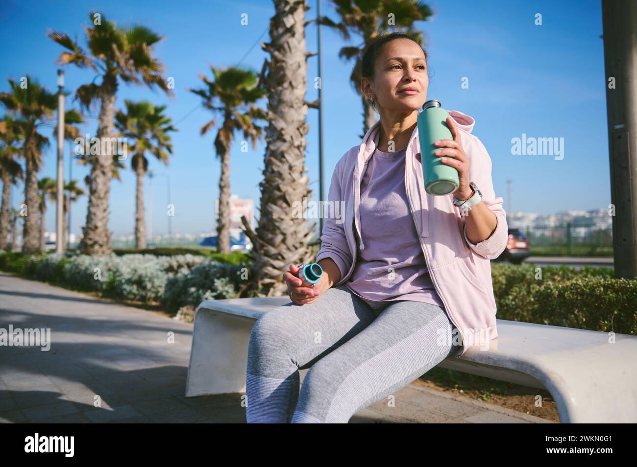 Attractive sporty woman in activewear, sitting on a bench on the marine promenade, drinking refreshing water after workout or jogging, relaxing and dr Stock Photo