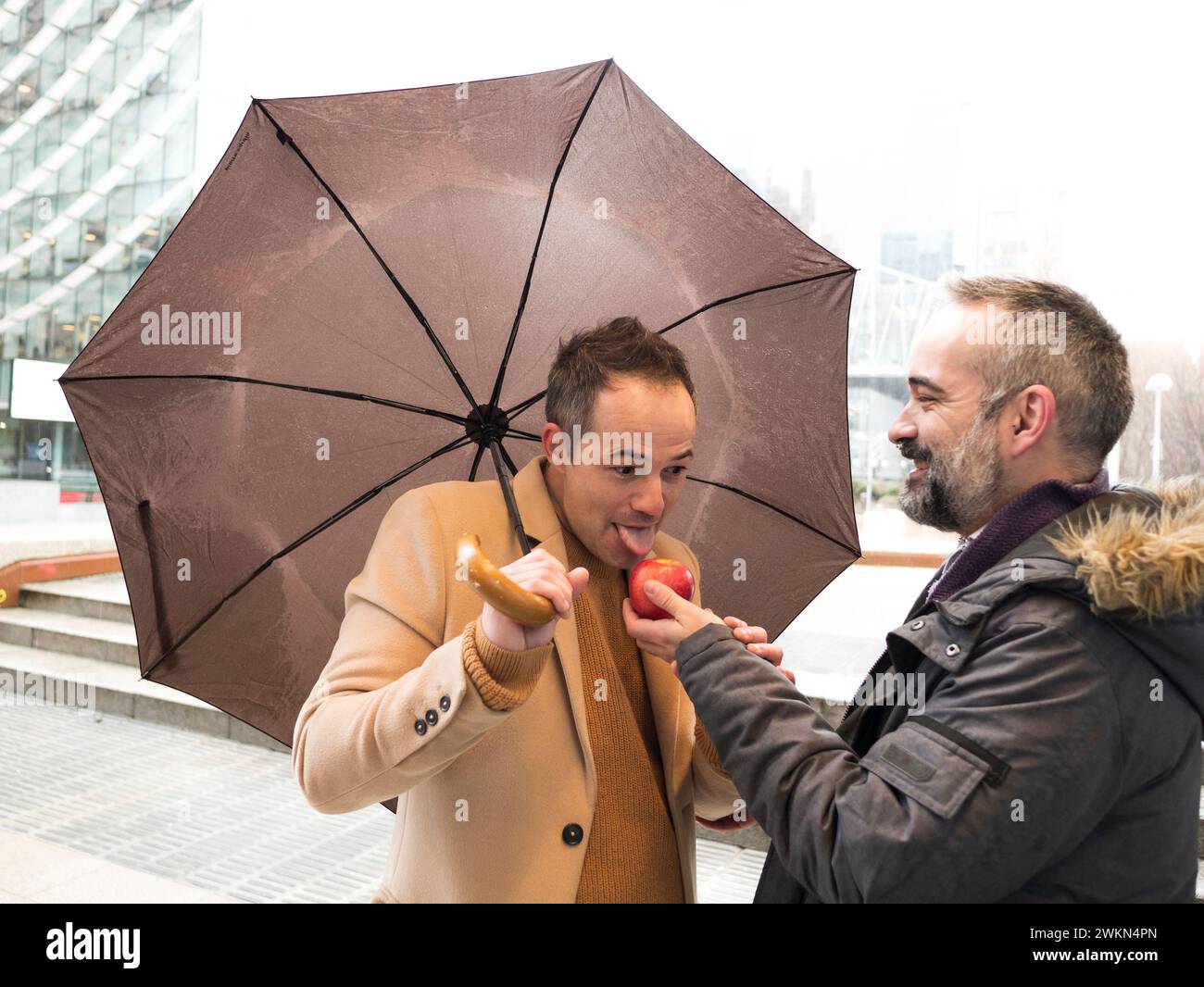 An executive offers a red apple to another who sucks it under the umbrella on a rainy day Stock Photo