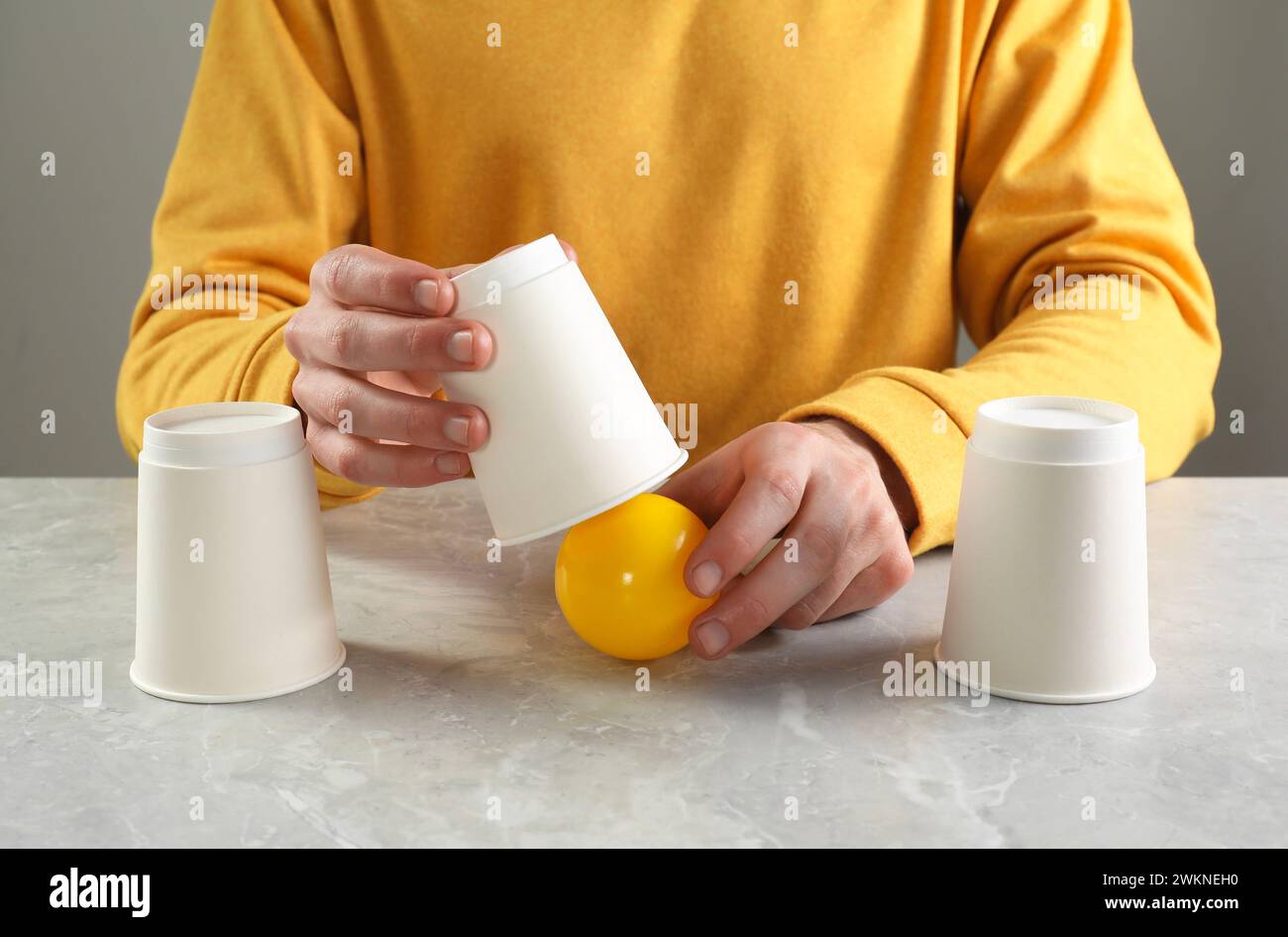 Shell game. Man showing ball under cup at light marble table, closeup Stock Photo