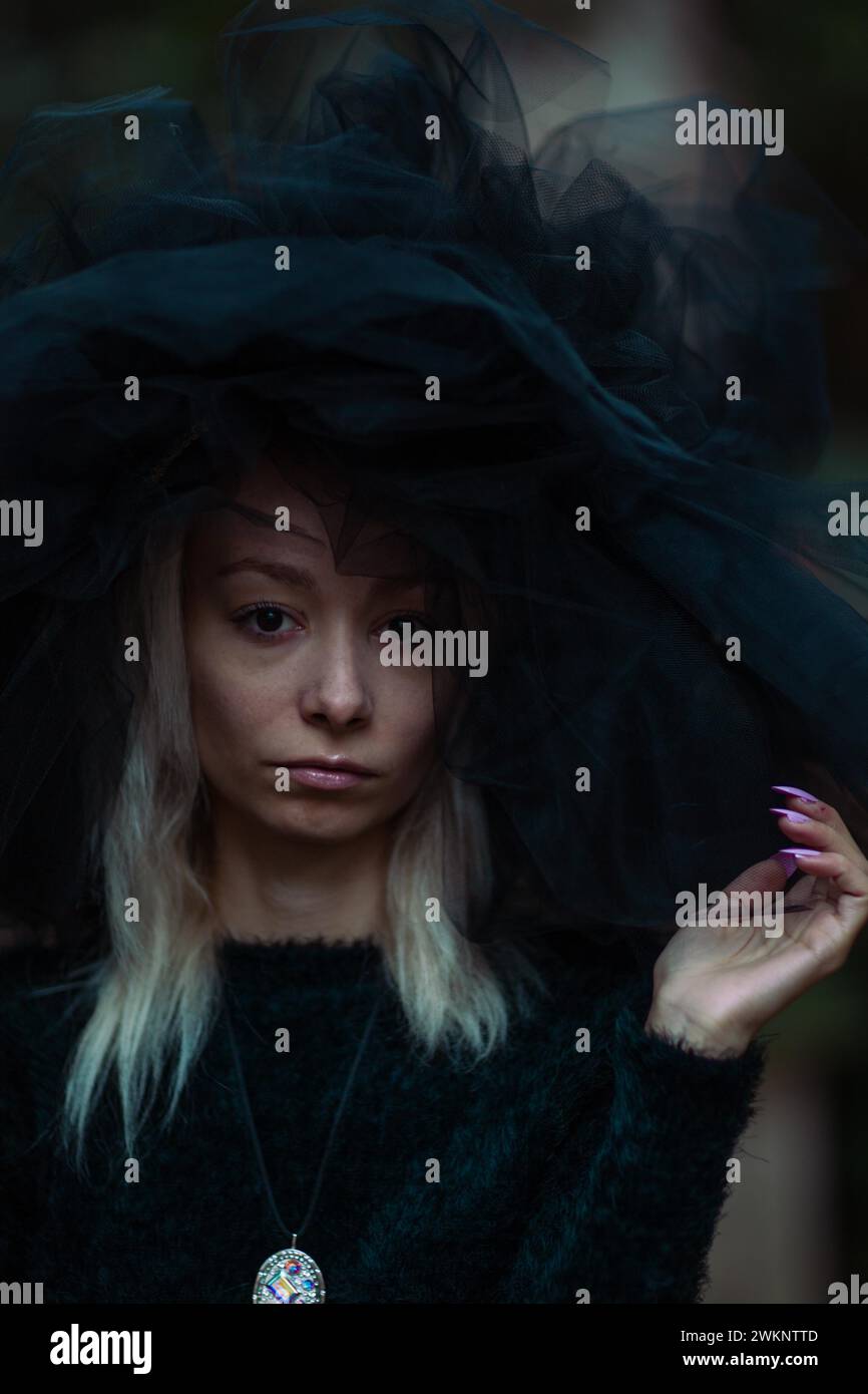 Mysterious white young blonde woman in a dark setting wear a large black tulle hat and veil and a black furry dress Stock Photo