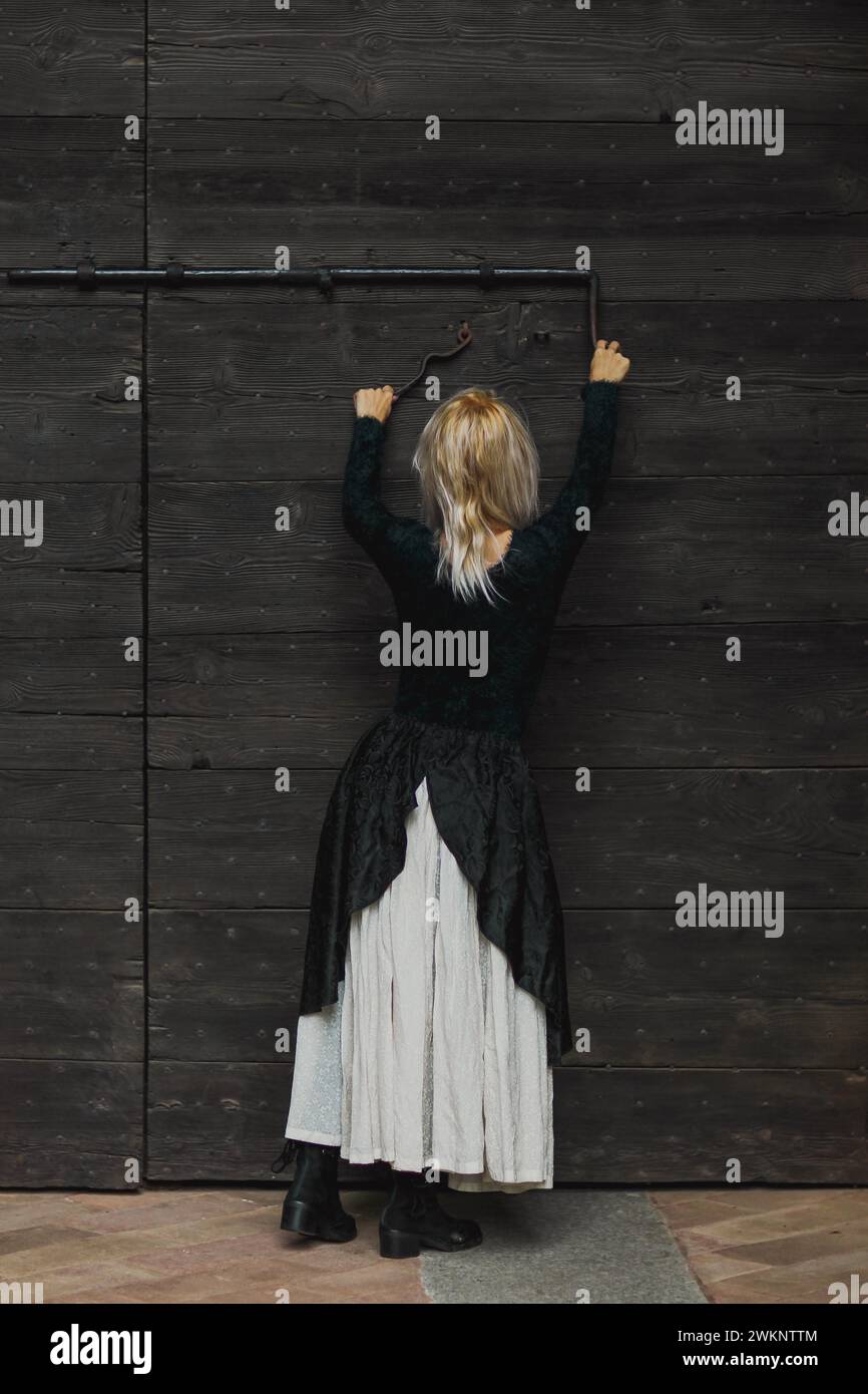 Full body shot of a young blonde caucasian woman reaching an antique lock of a wooden door above her head, wearing a black top and white skirt and a Stock Photo