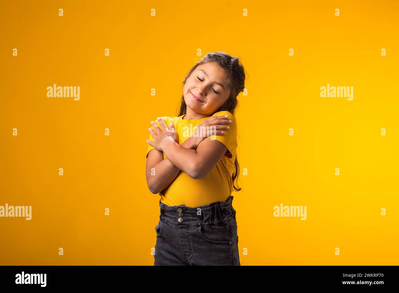 Portrait of child girl standing over isolated yellow background hugging oneself happy and positive, smiling confident. Self love and self care Stock Photo