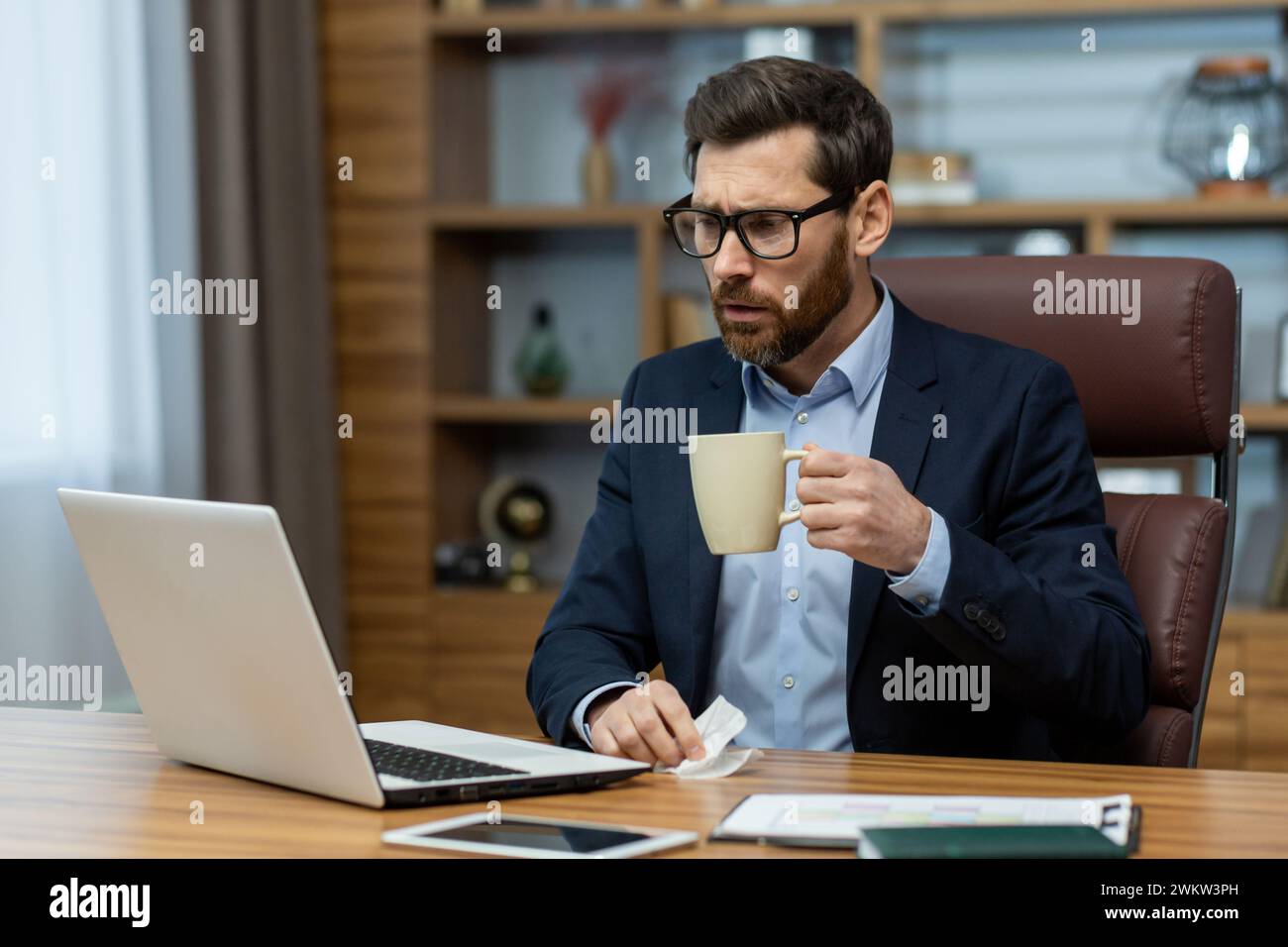 Serious and shocked young man working in the office, sitting at the desk, holding a cup with a drink in his hand and looking worriedly at the laptop s Stock Photo