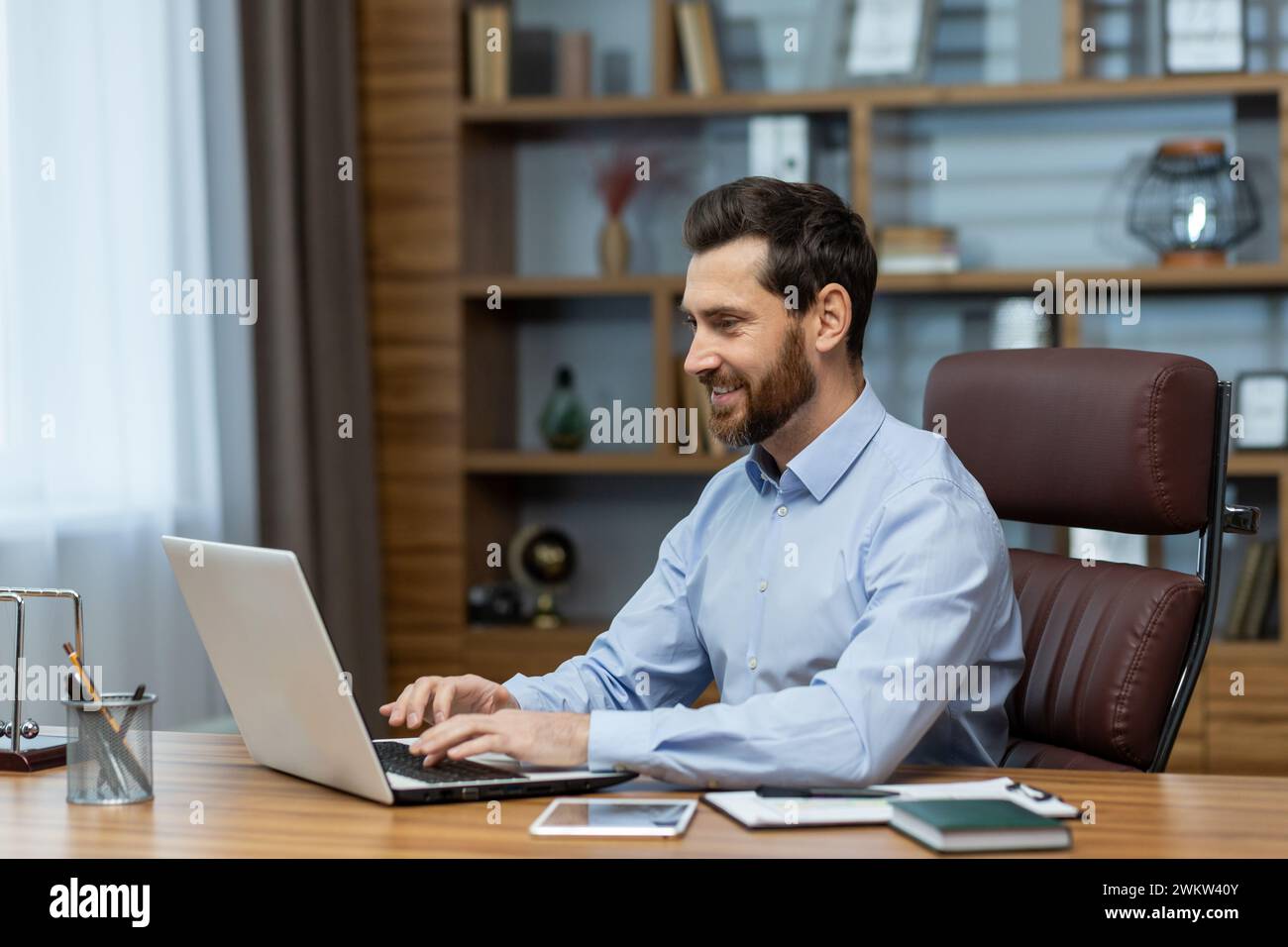 A smiling young man is working in the office, sitting at a laptop and typing on the keyboard. Stock Photo
