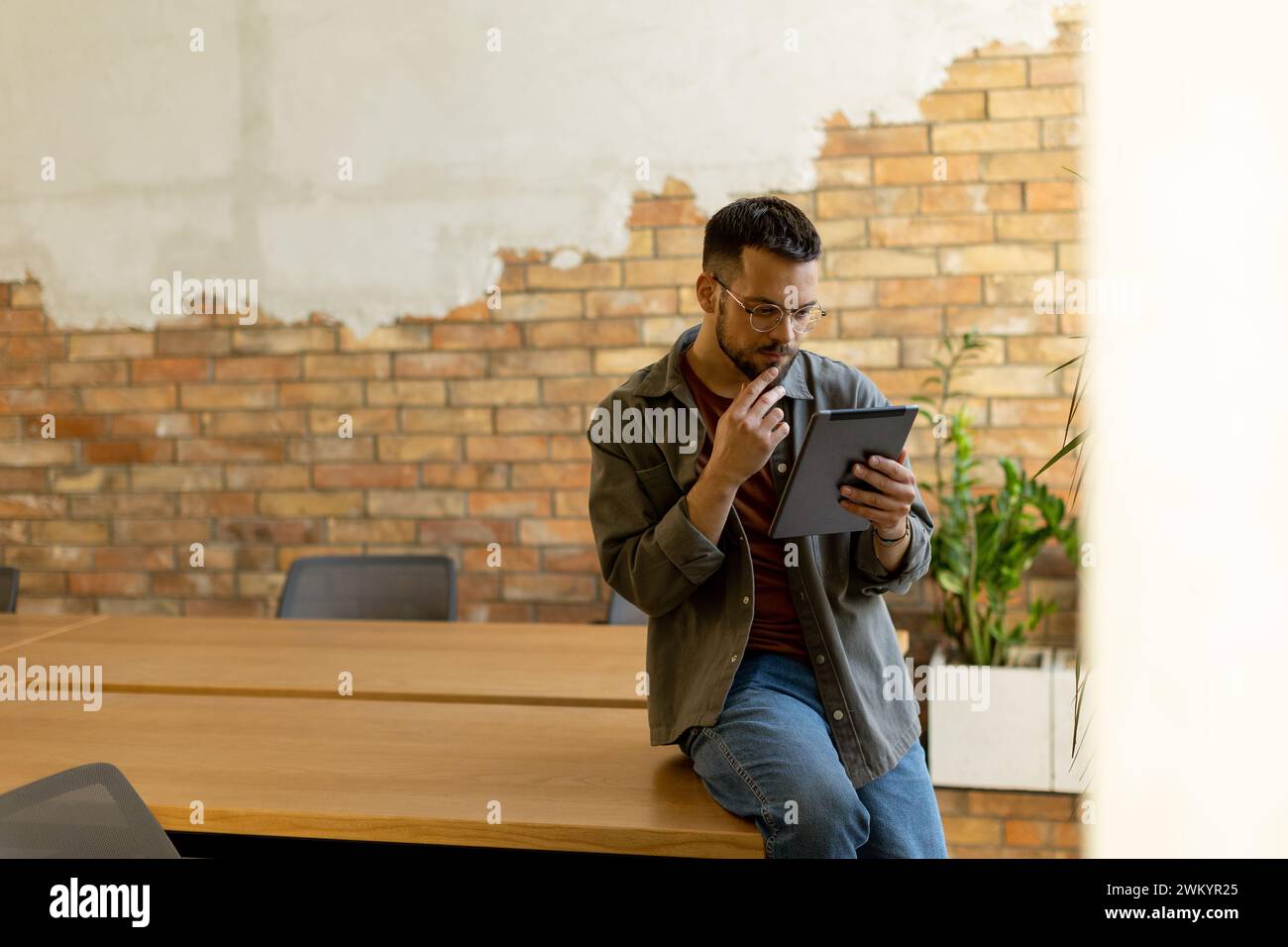 Cheerful man stands confidently holding a digital tablet in a contemporary office space with an exposed brick wall, symbolizing a blend of modern tech Stock Photo