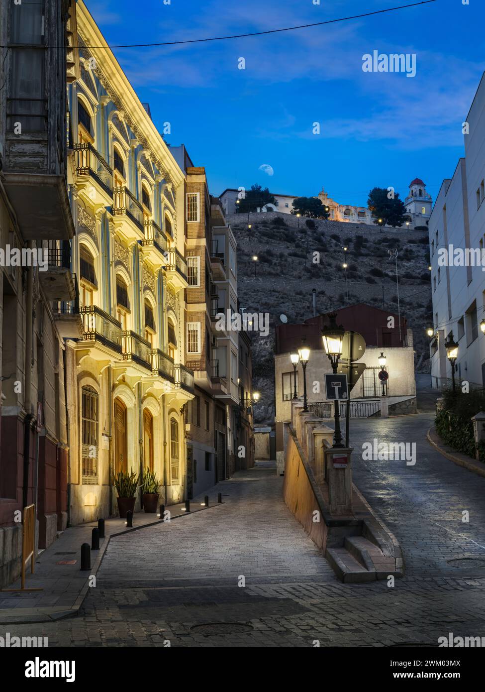 The streetlights flicker on as the moon rises above a deserted street in Orihuela, Alicante, Spain. Stock Photo
