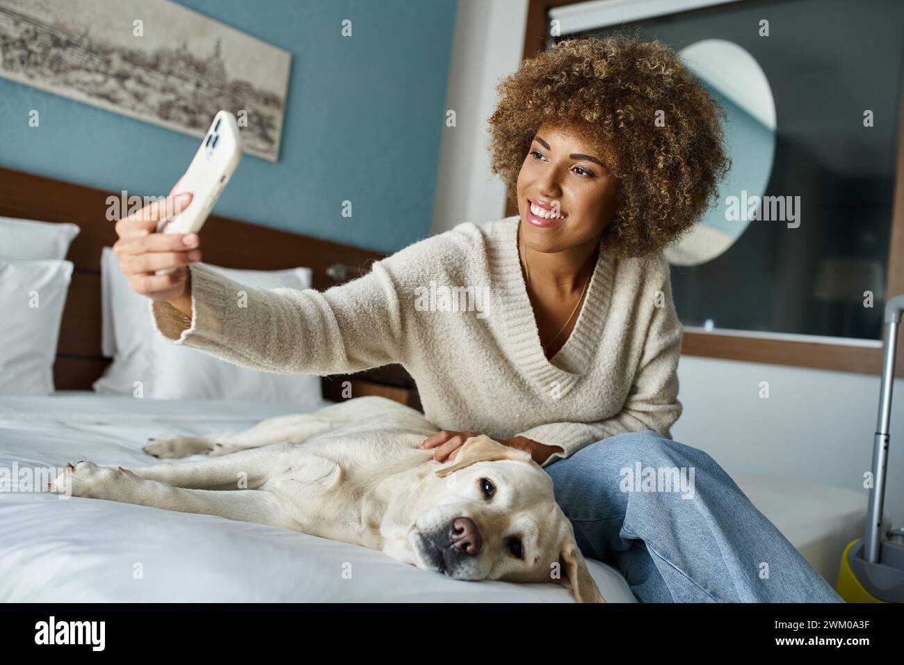 happy african american woman taking selfie with labrador on a bed in a