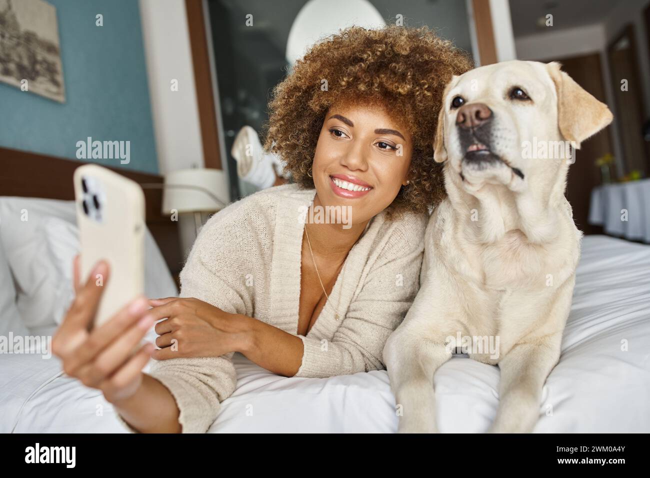 positive african american woman taking selfie with labrador on a bed in