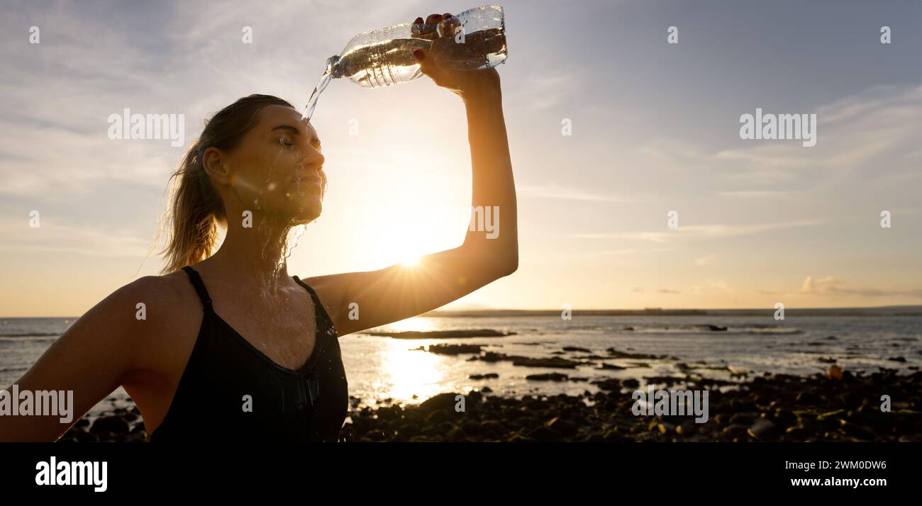 sporty woman pouring refreshing water on face from bottle after outdoor fitness workout on beach Stock Photo