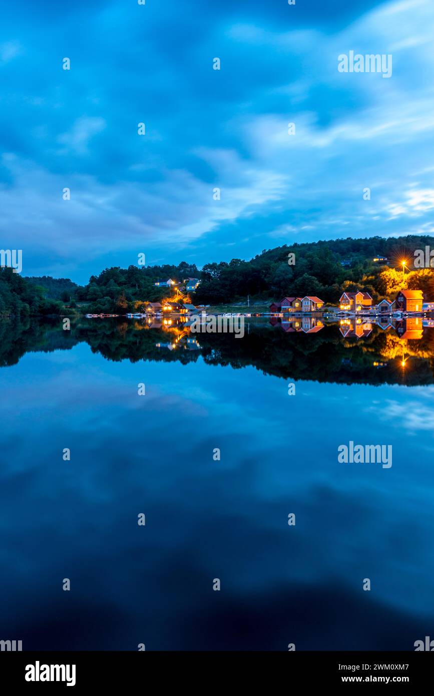 In the evening, the lights of the shore development are reflected in a mirror-smooth lake under a blue, spectacularly cloudy sky. Stock Photo