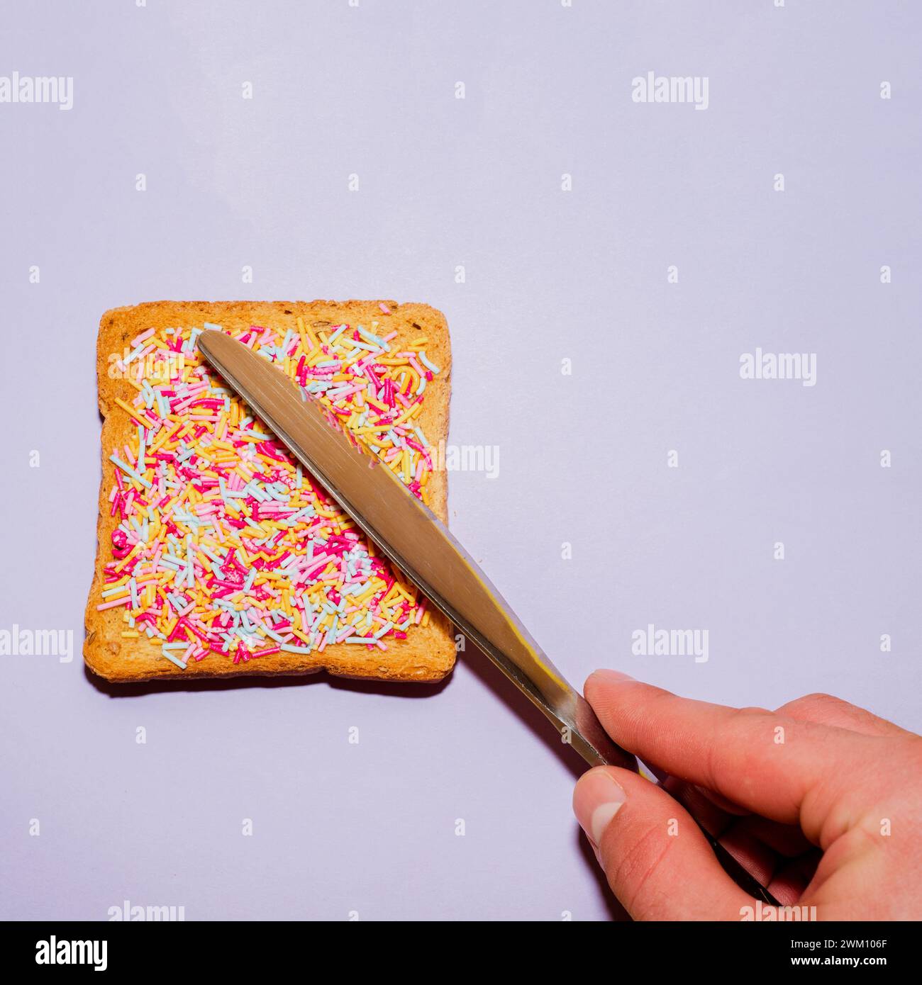 Knife spread colorful sprinkles on toast on a purple background. Creative food concept. Stock Photo