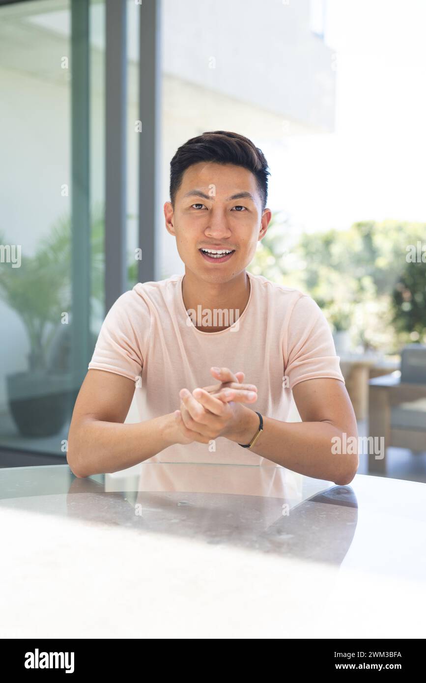 Young Asian man sits at a table, smiling confidently on a video call Stock Photo