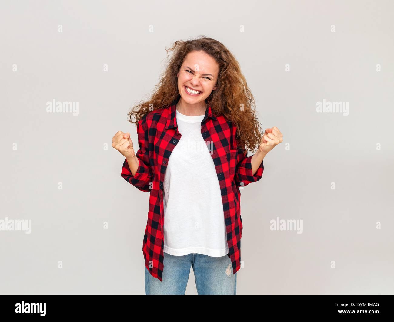 Young adult woman with brown hair  wearing red casual shirt rejoices in front of light grey coloured wall. Stock Photo