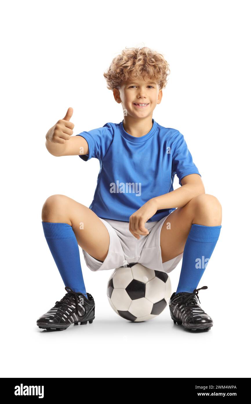 Happy boy in a sport jersey sitting on a football and gesturing thumbs up isolated on white background Stock Photo