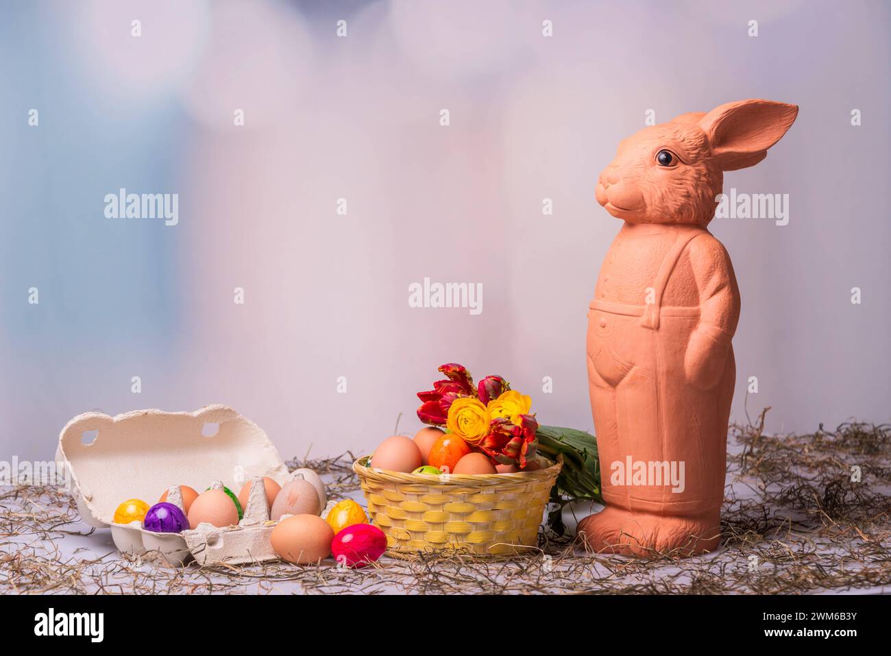 Eine Osterhasenfigur aus Ton vor einem Korb mit Ostereiern und buntem Hintergrund *** A clay Easter bunny figurine in front of a basket with Easter eggs and a colorful background Stock Photo