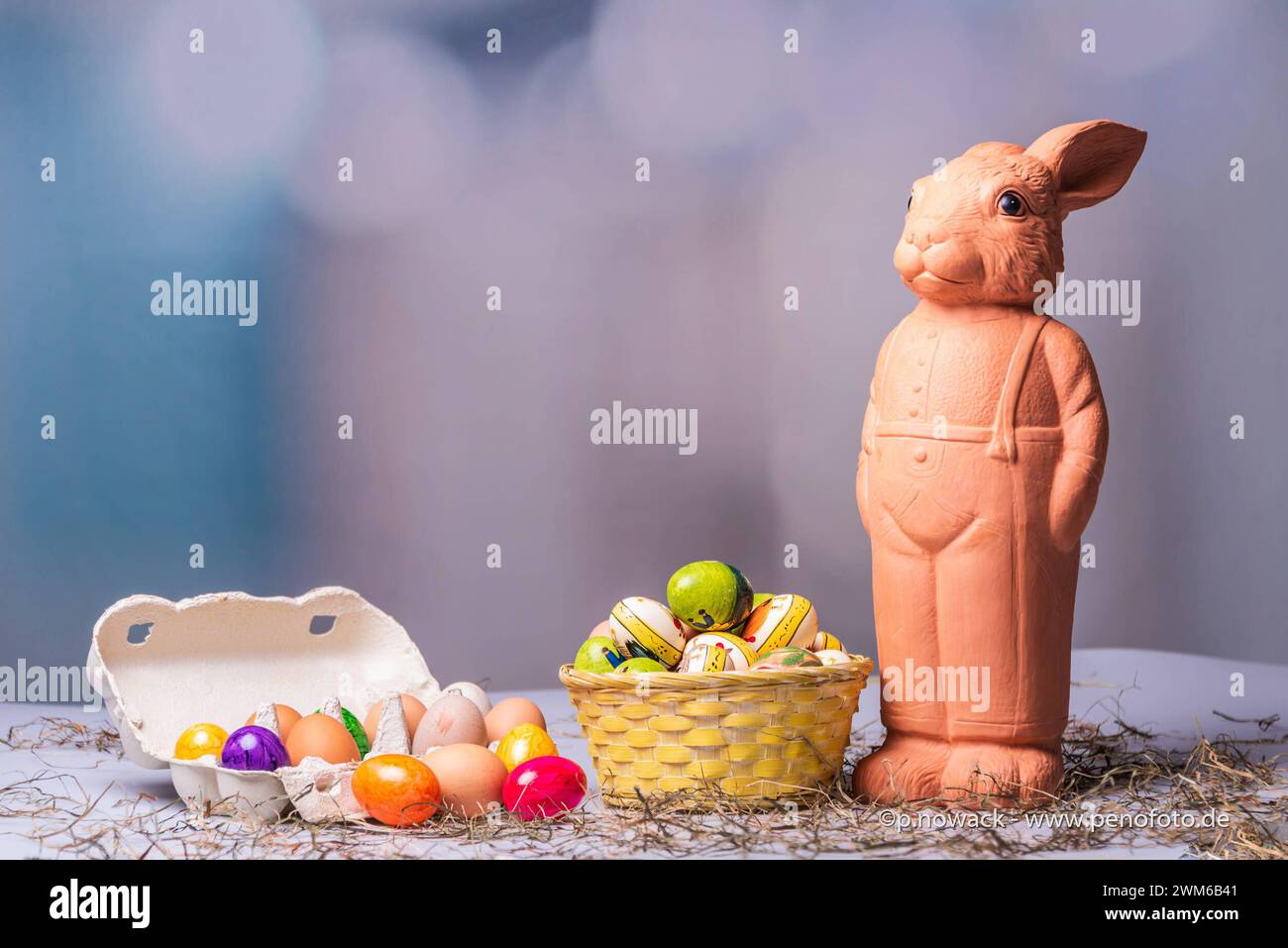 Eine Osterhasenfigur aus Ton vor einem Korb mit Ostereiern und buntem Hintergrund *** A clay Easter bunny figurine in front of a basket with Easter eggs and a colorful background Stock Photo