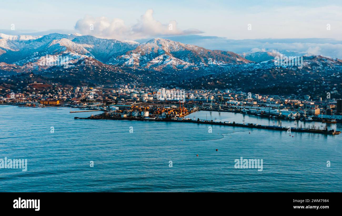Seaport on the coast against the background of mountains. A coastal city basks in the golden light of dusk, nestled between the calm sea and a dramatic backdrop of snow-capped mountains. The urban landscape merges seamlessly with the grandeur of nature. Stock Photo
