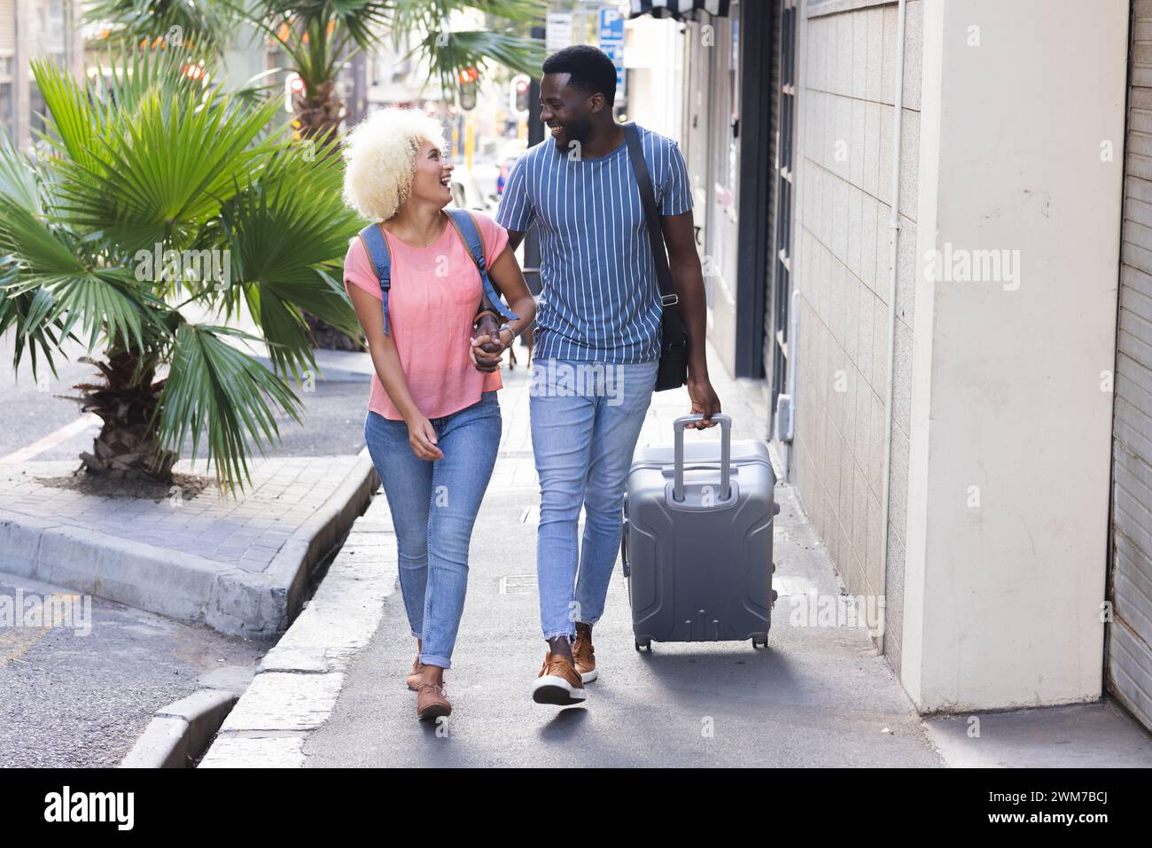 A young diverse biracial couple walks together in a city carrying luggage on vacation Stock Photo