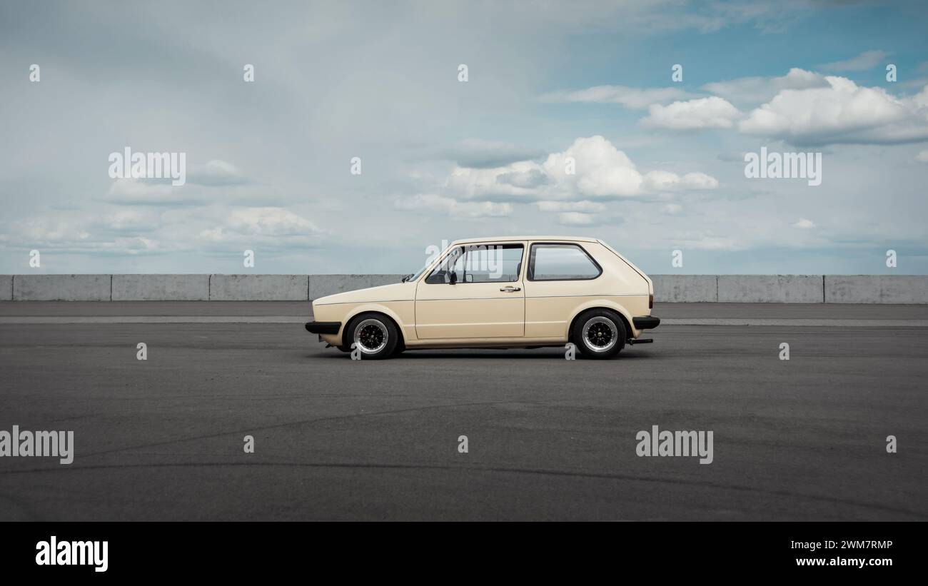 Side profile view of classic Volkswagen Golf hatchback. Beige Volkswagen Golf mk1 standing on big empty parking lot with cloudy sky in the background. Stock Photo