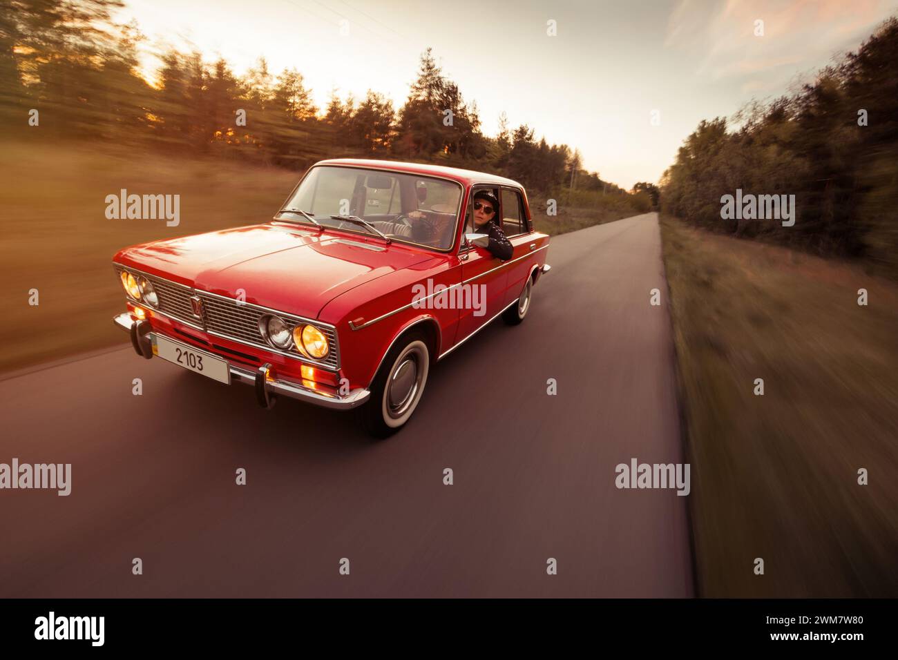 classic VAZ 2103 sedan from 1970s driving on narrow road. Three quarter front view motion shot of red Lada at sunset. Stock Photo