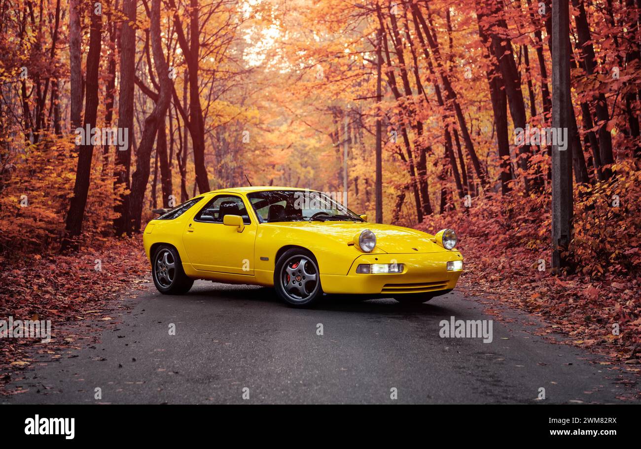 yellow Porsche 928 in autumn forest. Three quarter front view of 1980s german sportscar on narrow road in deciduous forest. Stock Photo