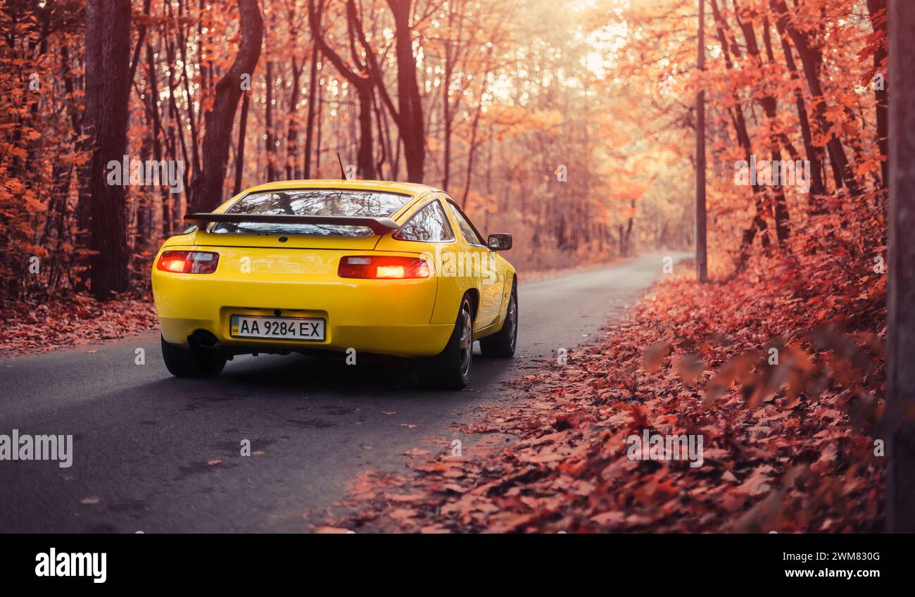 yellow Porsche 928 in autumn forest. Three quarter rear view of 1980s german sportscar on narrow road in deciduous forest. Stock Photo