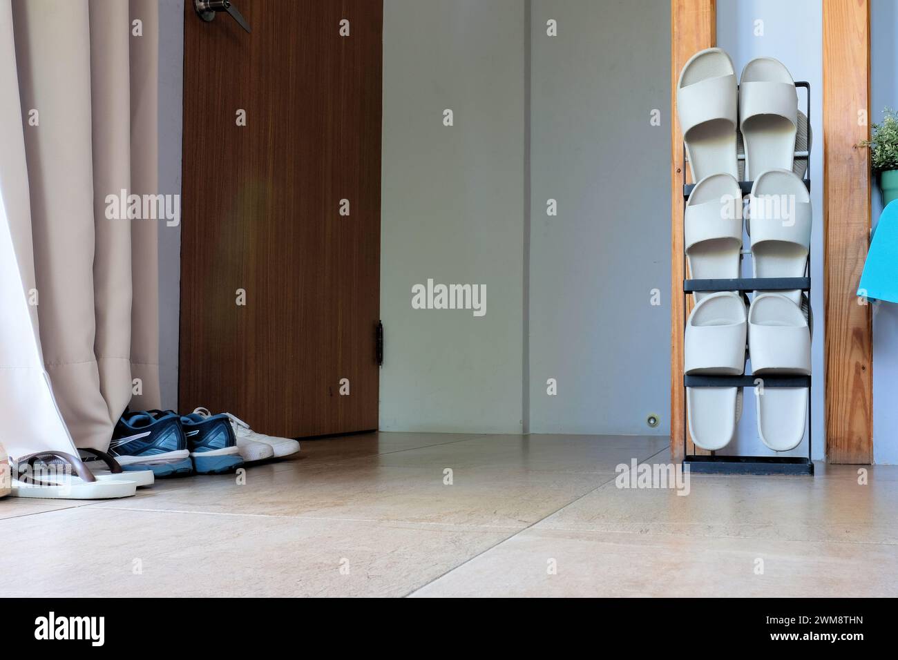 Vertical shoe rack holding three pairs of rubber sandals for use indoors located next to door and entryway with outdoor shoes on floor; Asia, Taiwan. Stock Photo