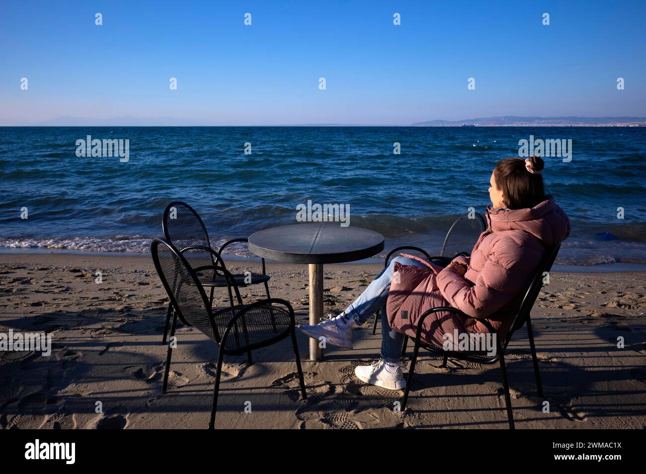 Young woman looking at the sea, lonely, alone, coat, table and chairs, beach bar, beach, Peraia, also Perea, Thessaloniki, Macedonia, Greece Stock Photo