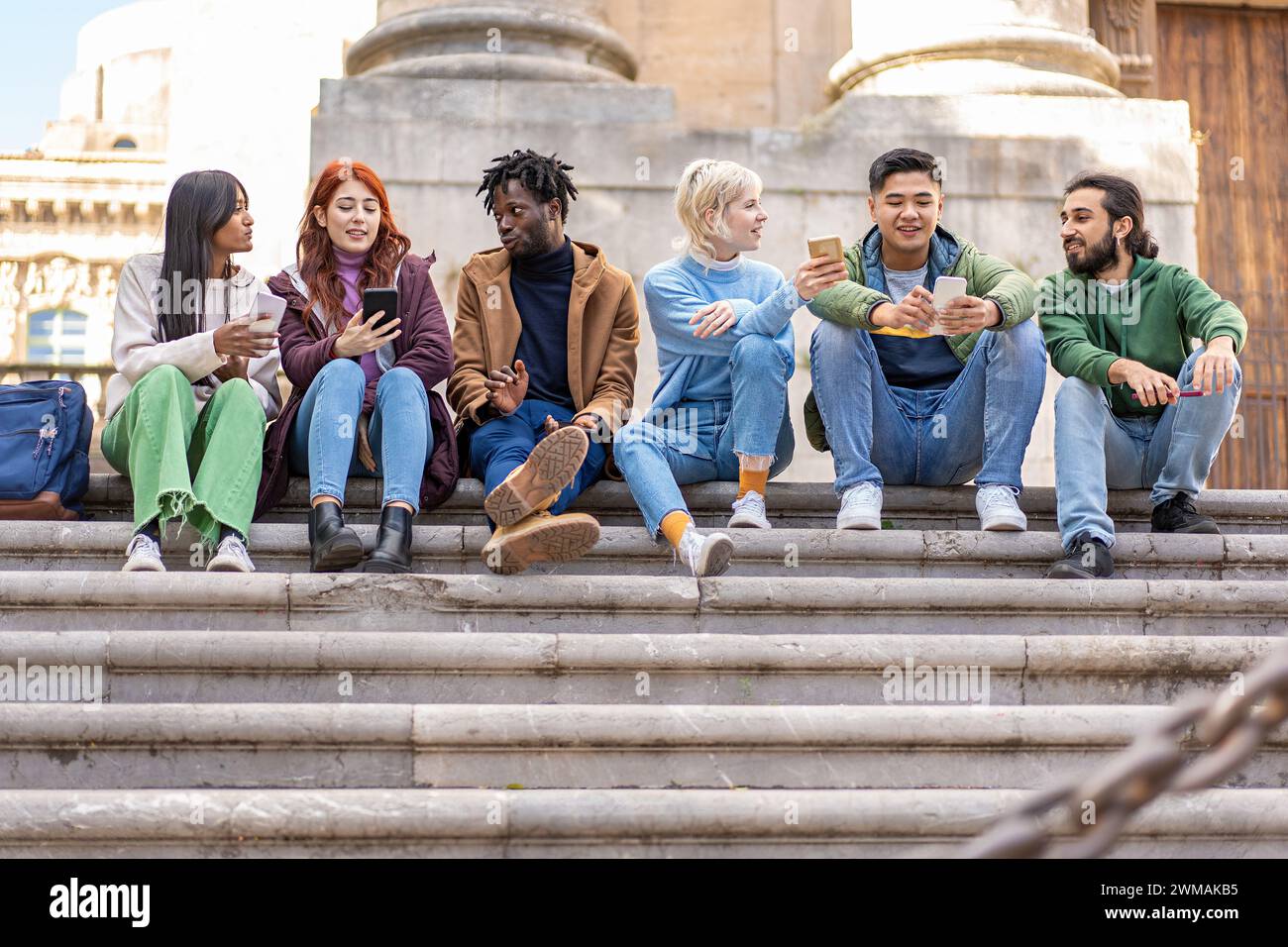 Group of young adults engaged in conversation while sharing content on smartphones, reflecting a relaxed urban lifestyle. Stock Photo