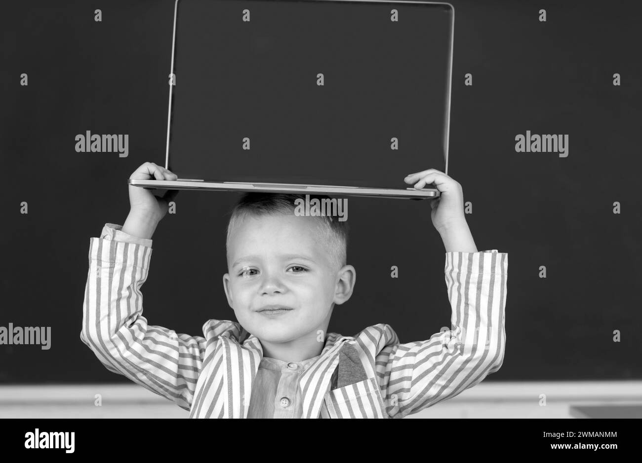 School child holding laptop on head using digital pc laptop in class. Portrait of funny pupil of primary school study programming. Stock Photo