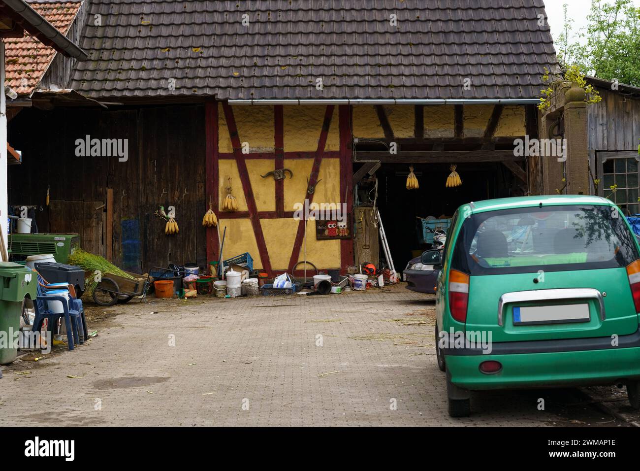 A green car is parked in front of an open rustic farmhouse garage revealing a cluttered interior, with various tools, buckets, and equipment scattered Stock Photo