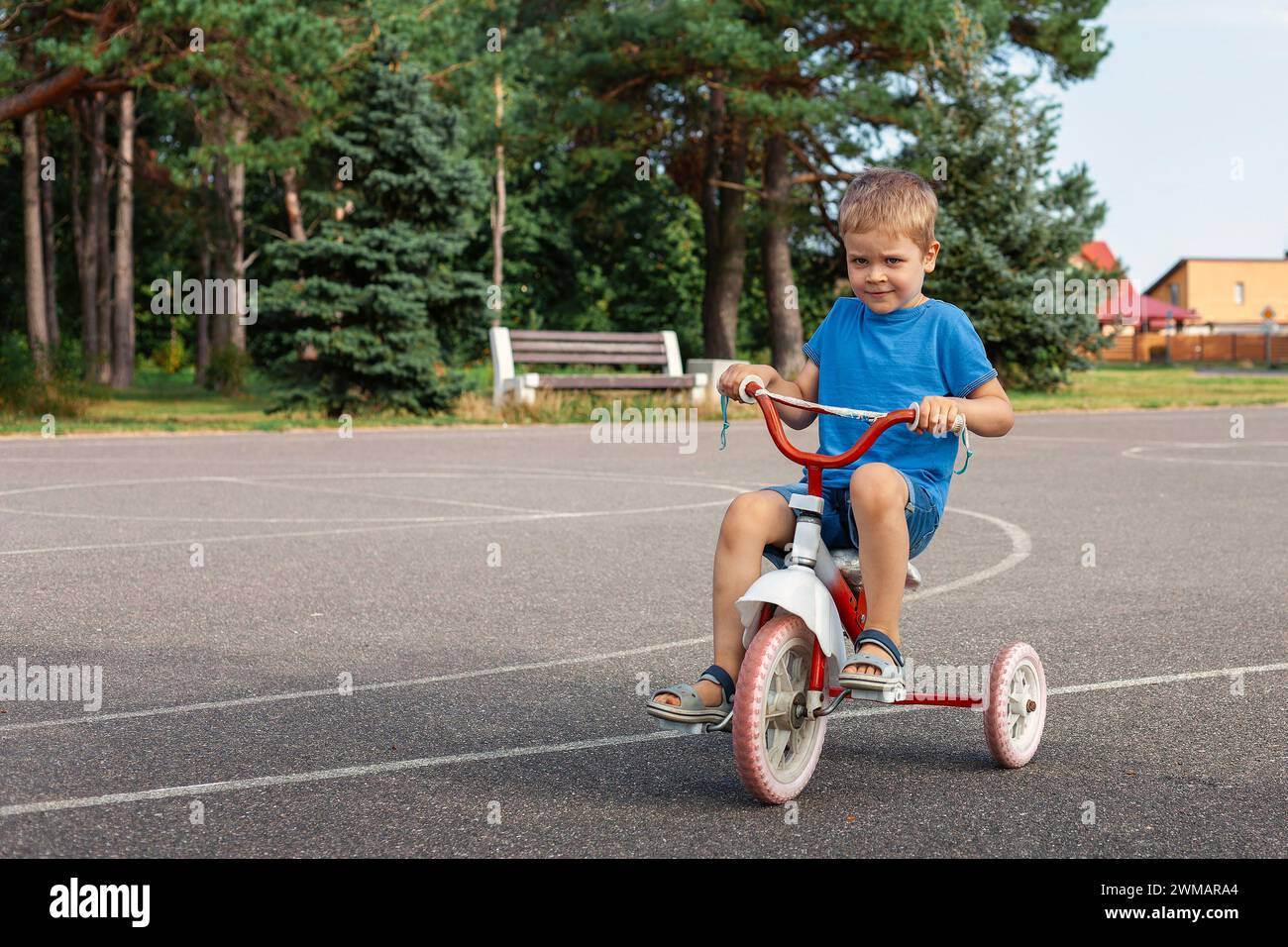 Smiling cute boy in blue clothes is riding a tricycle in a park lot. Stock Photo