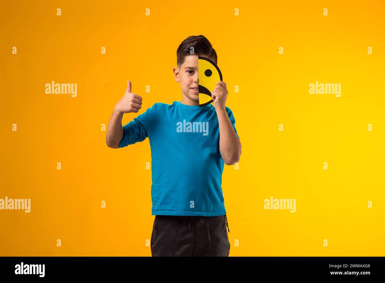Portrait of child boy holds emoticons on half face with sad emotions and shoing thumb up gesture over yellow background. Stock Photo