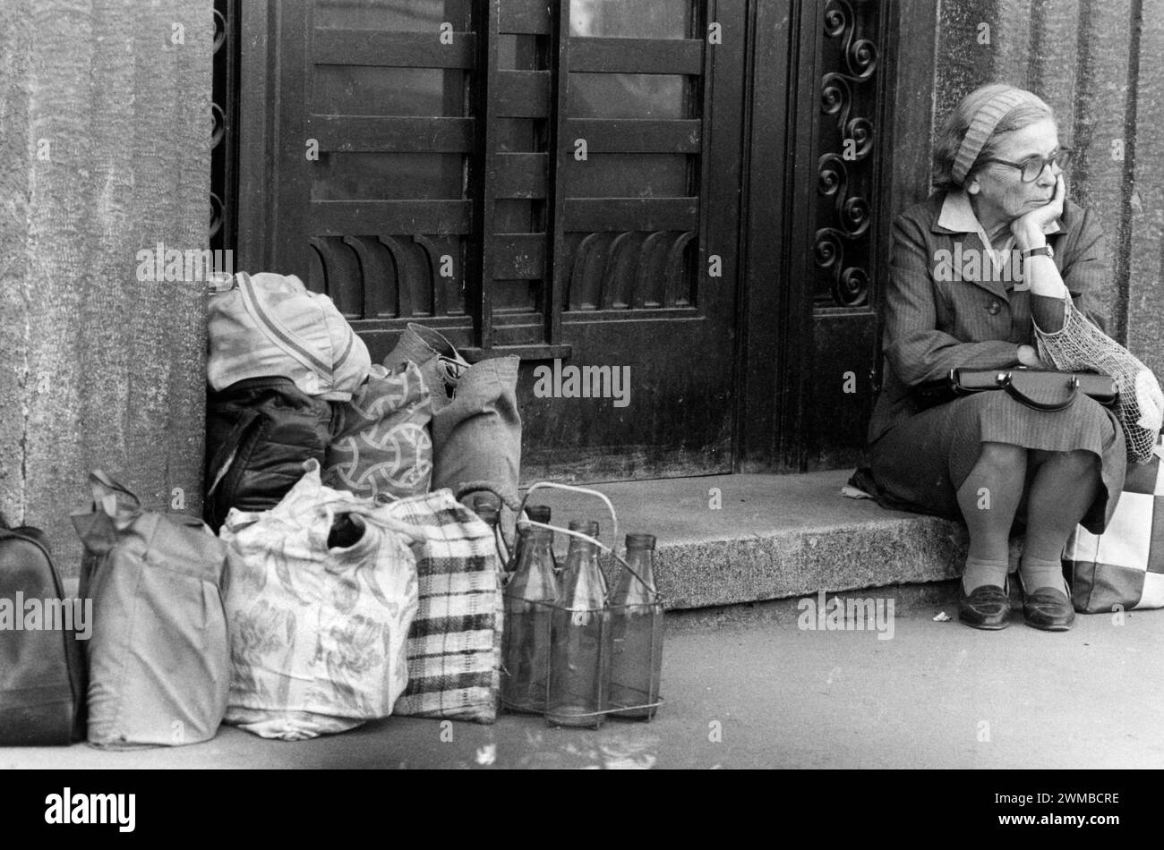 Bucharest, Romania, February 1990. Shortly after the anti-communist revolution of December 1989, people still stay in long lines to get hold of basic groceries. The centralized socialist economic system created scarcity and hunger. Here, an elderly woman is sitting down, waiting in line for the delivery of milk. Other people left their bags with reusable glass bottles, reserving their spot in line. Stock Photo