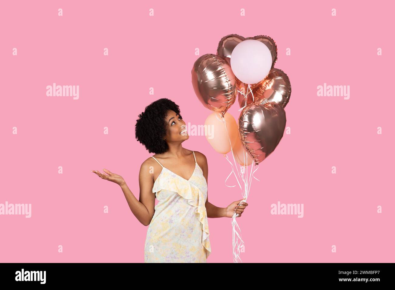 Cheerful African American woman with curly hair, looking up at rose gold and white balloons in her hand Stock Photo