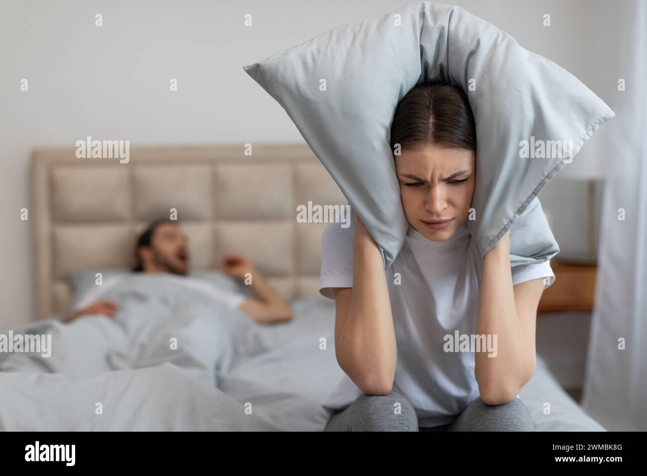 Annoyed woman covers ears with pillow to block snoring Stock Photo